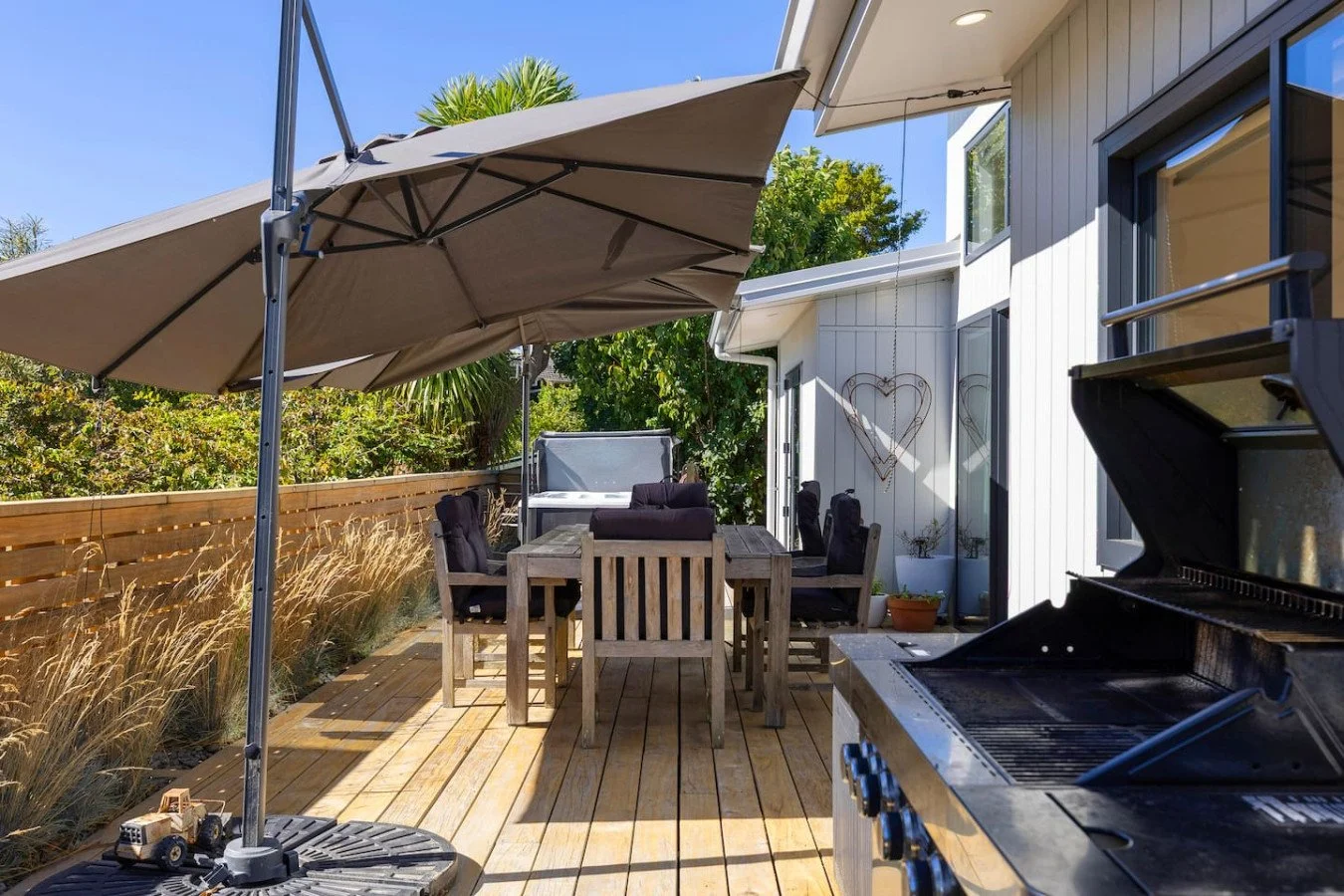Backyard patio with wooden deck, large umbrella, outdoor dining table with six chairs, a grill, and decorative heart-shaped wall art in front of a white house.