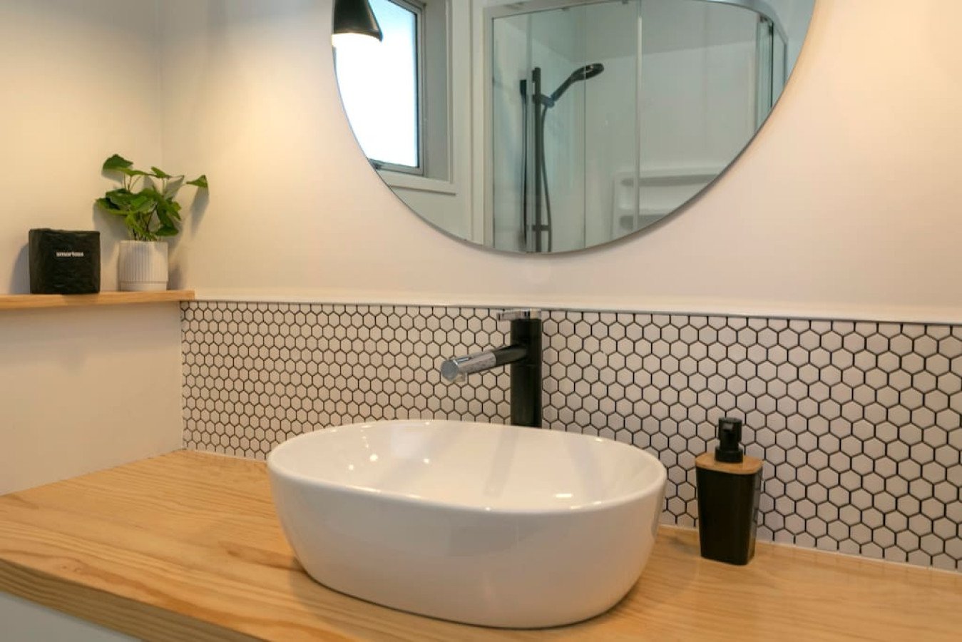 Bathroom sink with a wooden countertop, a white oval bowl, and a black soap dispenser. A large round mirror is above the sink, reflecting a shower with a handheld showerhead and a window.