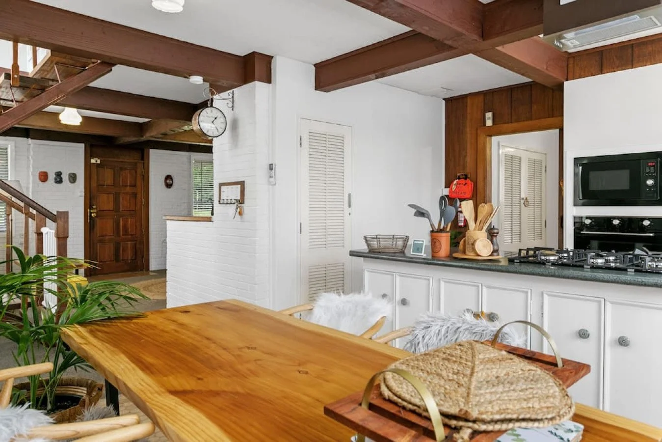 Inside a cozy kitchen and dining area with a wooden table, potted plant, and kitchen counter with utensils, microwave, and oven in the background. The space features wood accents and white walls.