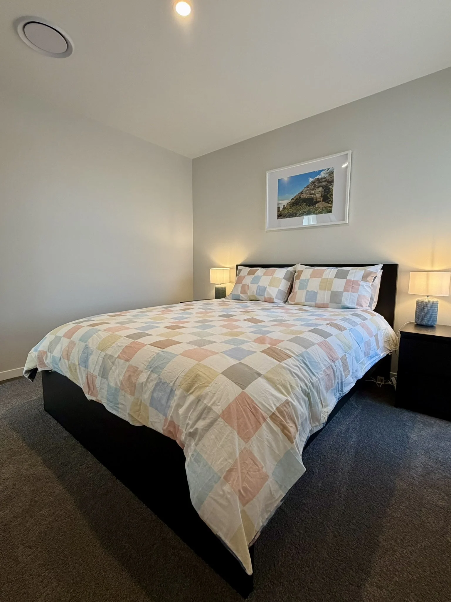 A neatly made bed with a pastel checkered quilt and matching pillows, flanked by two small nightstands with lamps in a modern, minimalistic bedroom.