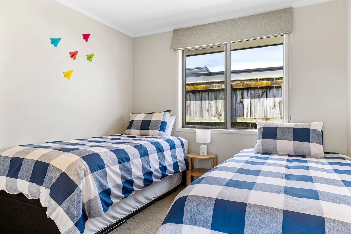 Two twin beds with blue and white checkered bedding, a small wooden nightstand with a lamp, a window showing a wooden fence outside, and colorful wall-mounted paper airplanes.