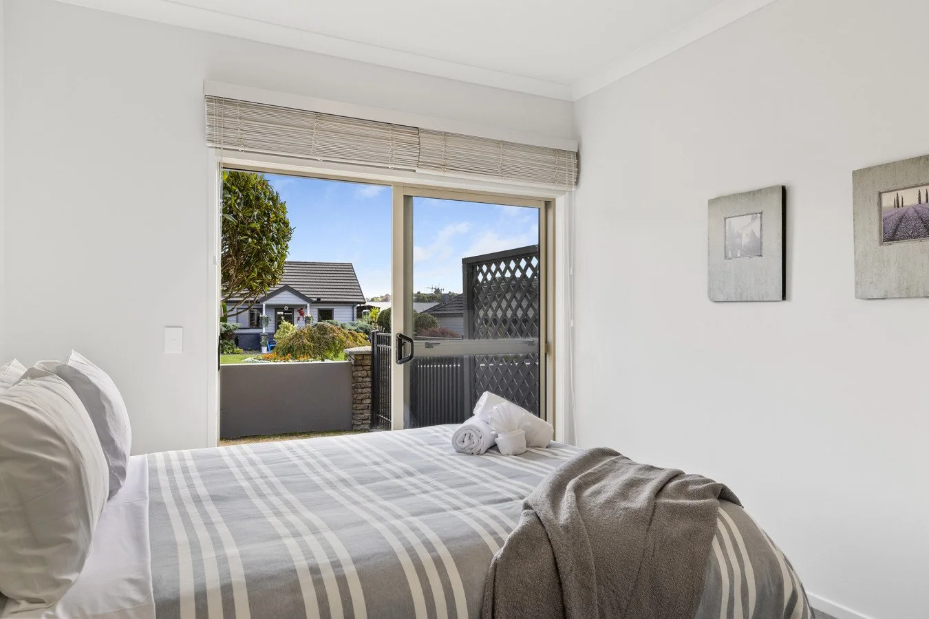 A bedroom with a bed that has white and beige striped bedding, pillows, and a folded towel. There is a sliding glass door opening to a small balcony with a view of neighboring houses and greenery. The room has white walls with two framed pictures han