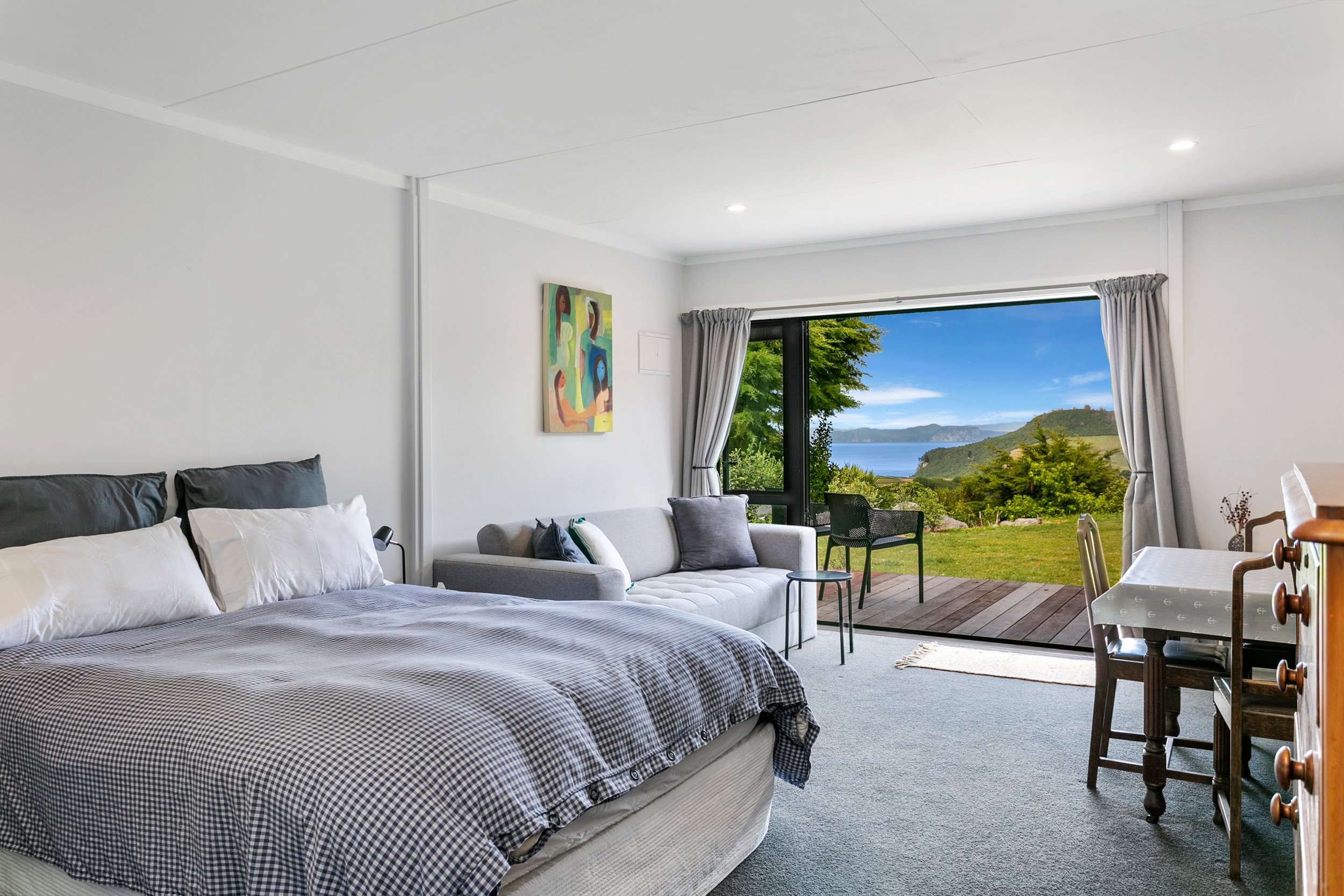 Room with a bed, gray and white bedding, a sofa with pillows, a small table, a dining table with chairs, a balcony overlooking green hills and water, with trees and a blue sky.