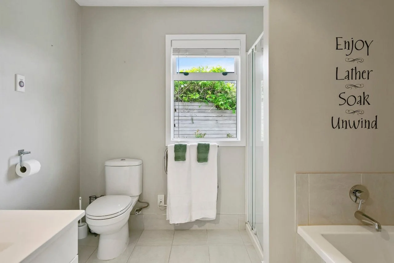 Bathroom with toilet, window, towel rack, shower, bathtub, and wall decal with words: "Enjoy, Lather, Soak, Unwind."