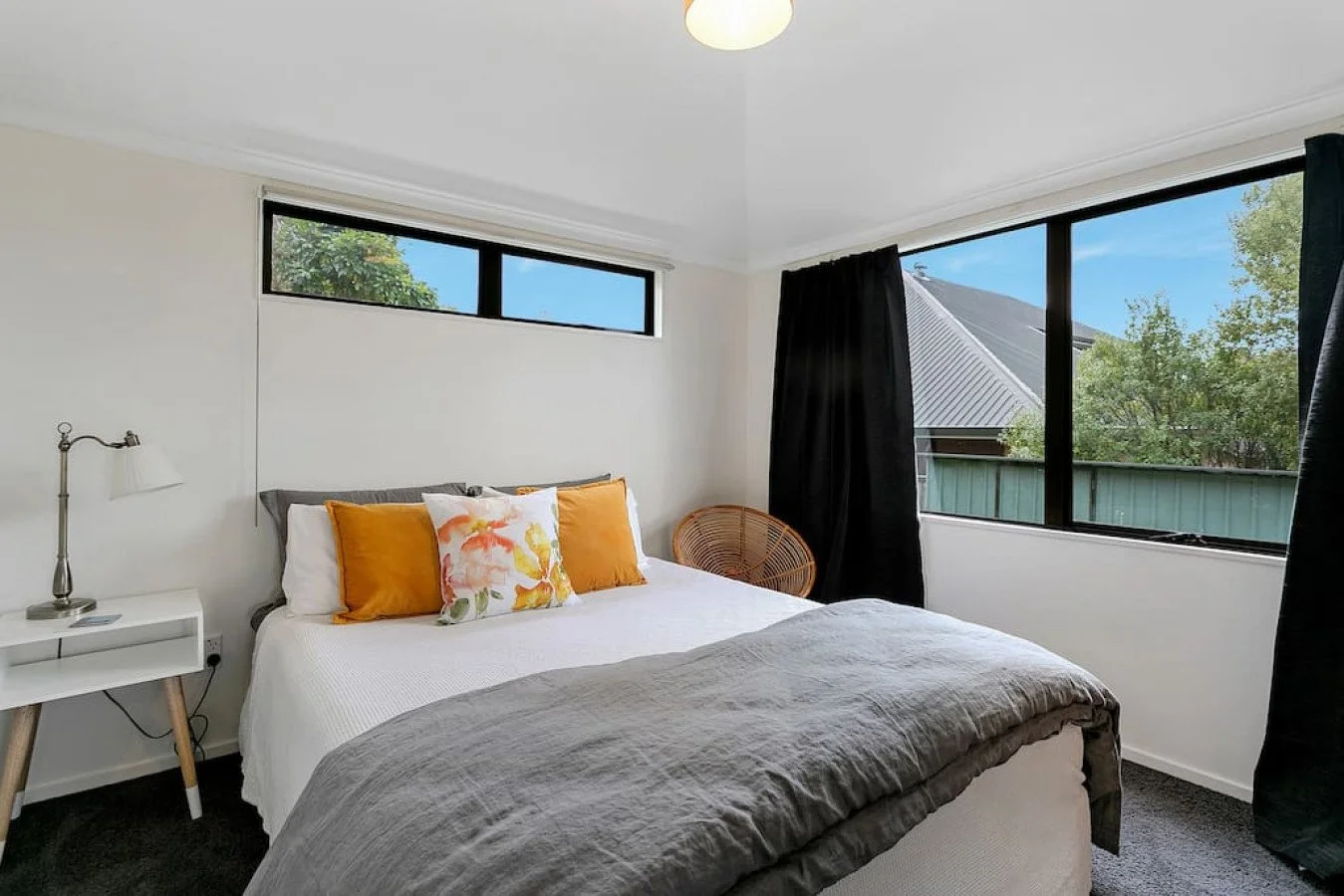 A bedroom with a large bed, gray and white bedding, and orange and floral pillows. There is a white nightstand with a lamp on the left, and a wicker chair by the window. The room has two large windows with black curtains, showing trees and a neighbor