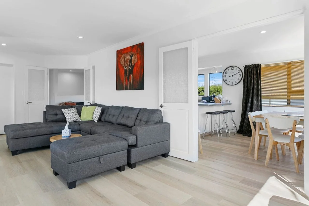 Modern living room with gray sectional sofa, colorful throw pillows, wooden coffee table, artwork of a dog on the wall, open kitchen with bar stools, round wall clock, and dining area with white table and chairs.