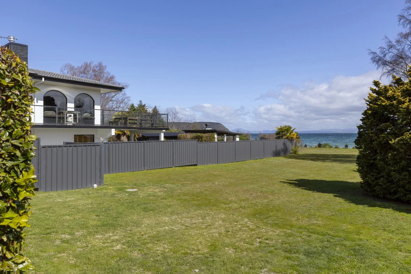 View of a lawn and a coastal house with a balcony, gray fence, shrubs, and ocean in the background on a partly cloudy day.