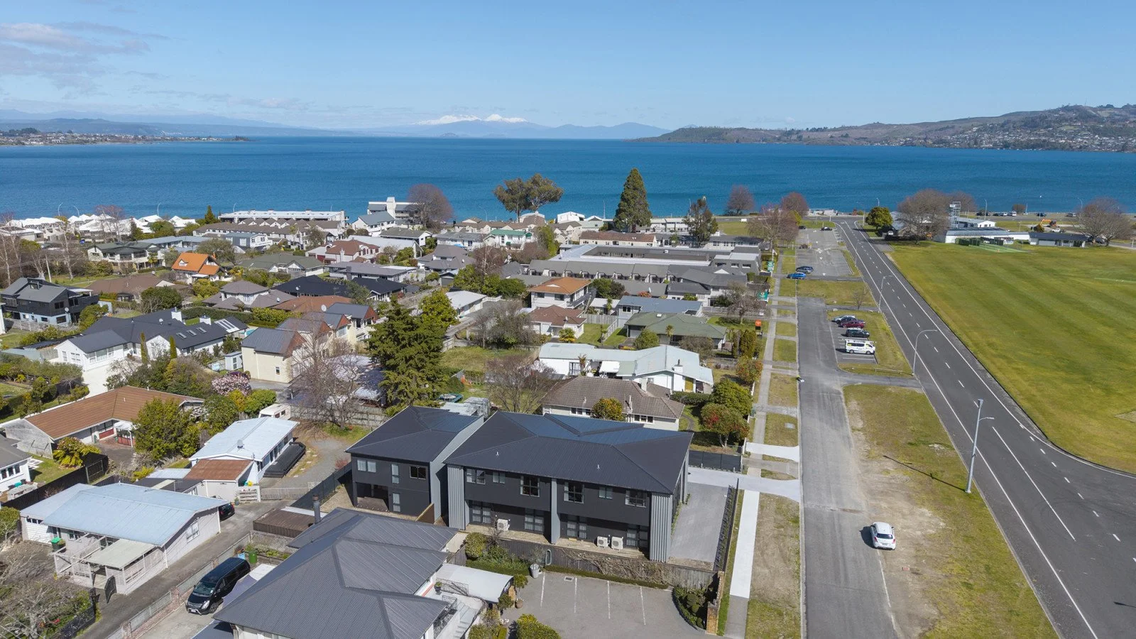 Aerial view of a coastal residential neighborhood with houses, a large green field, and a body of water in the background under a partly cloudy sky.