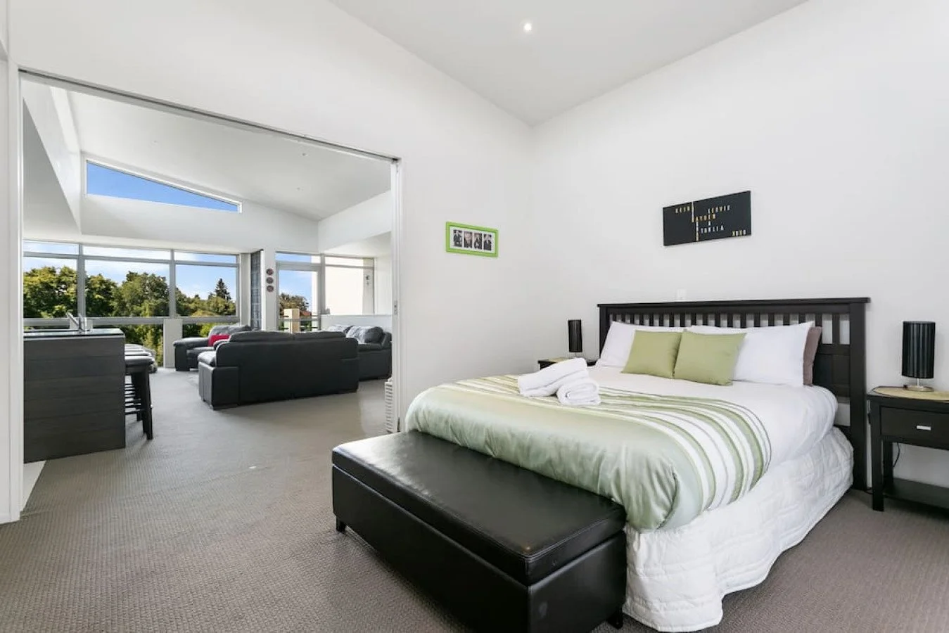 A bedroom with a king-size bed, pillows, and a bench at the foot of the bed. Two nightstands with lamps are on either side of the bed. A living area with sofas and large windows is visible in the background.