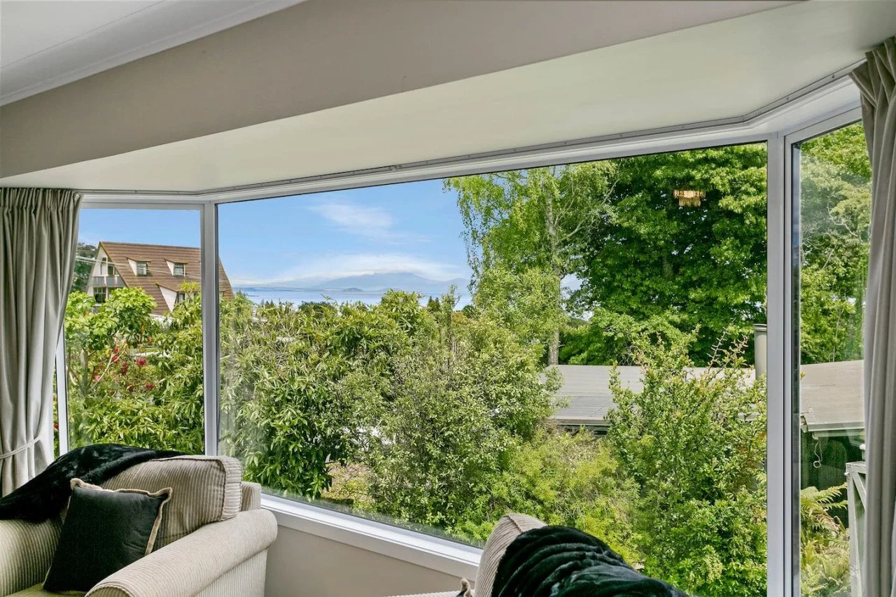 Interior view of a room with a large window showing a lush green garden and trees outside, with a glimpse of a neighboring house.