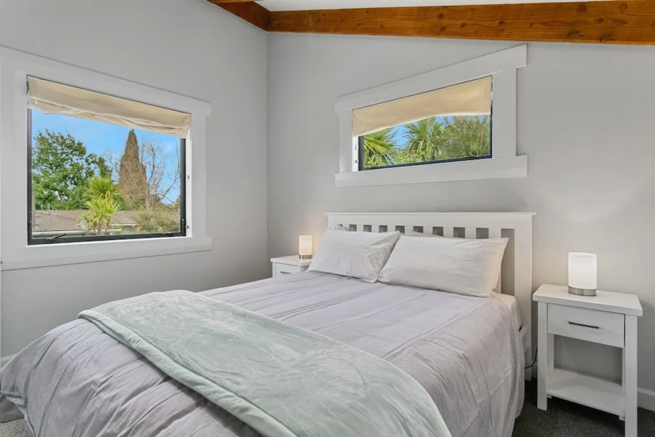 A bedroom with two windows, a white bed with pillows, a blanket, and two white nightstands with lamps, white walls, and a wooden ceiling.