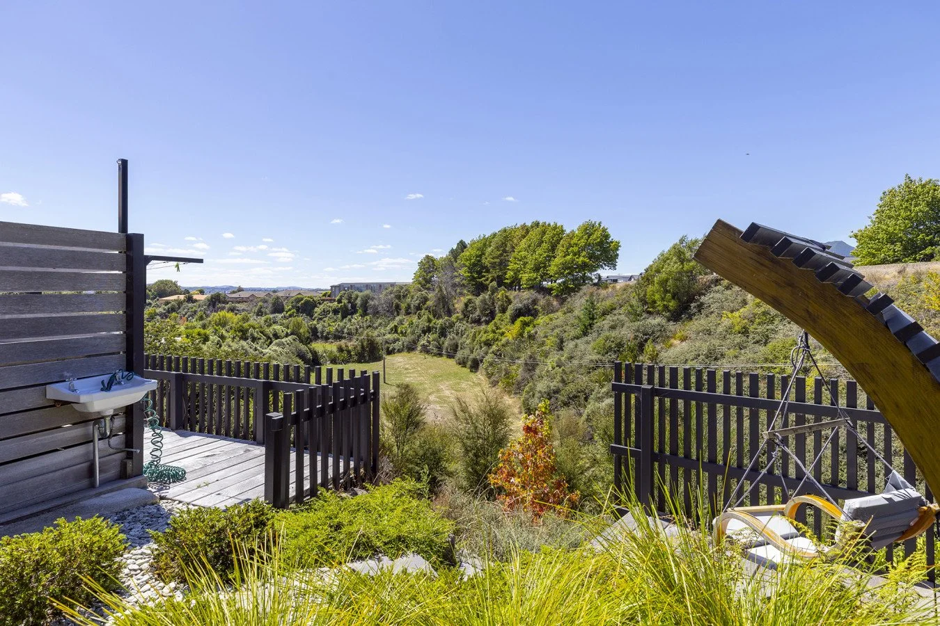 A backyard overlooking a green landscape with trees, shrubs, and a clear blue sky, featuring a black wooden fence, a wooden swing with a black canopy, and a small outdoor sink with a hose.