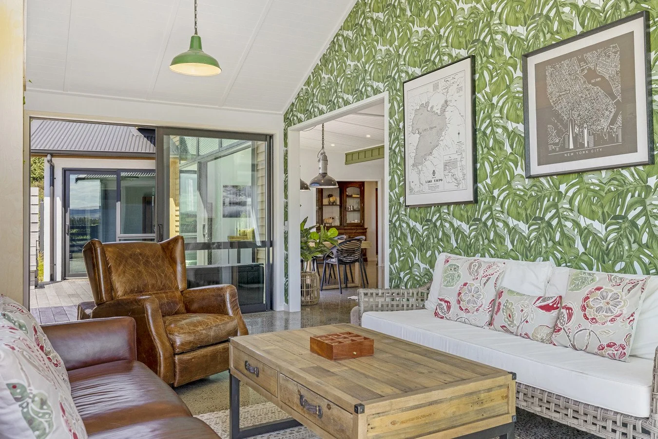 Living room with tropical leaf wallpaper, white sofa with floral pillows, brown leather armchair, wooden coffee table, and framed city maps on the wall, with an open door leading to an outdoor deck.