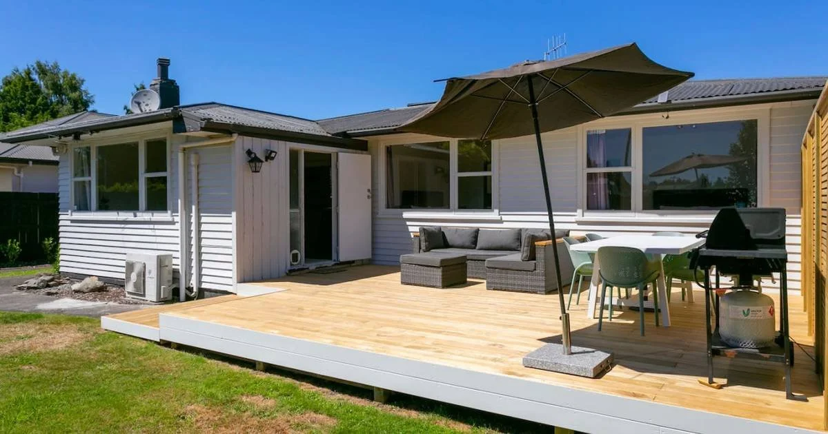 Backyard patio with outdoor furniture, table, chairs, umbrella, and grill in front of a house with white siding.