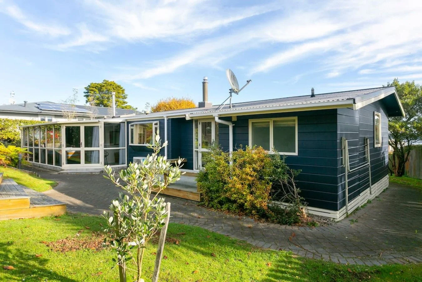 A blue house with a sunroom and solar panels on the roof, surrounded by a green lawn with trees and shrubs, under a blue sky with some clouds.
