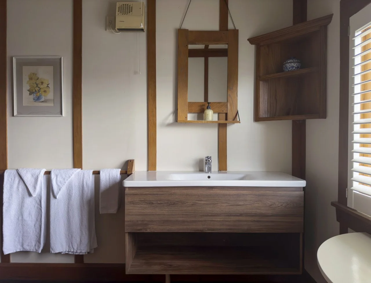 Bathroom with wooden accents, white towels hanging on a rack, sink with wooden cabinet, mirror, shelf with soap, picture of yellow flowers, window with blinds, and a wall-mounted heater or air conditioning unit.