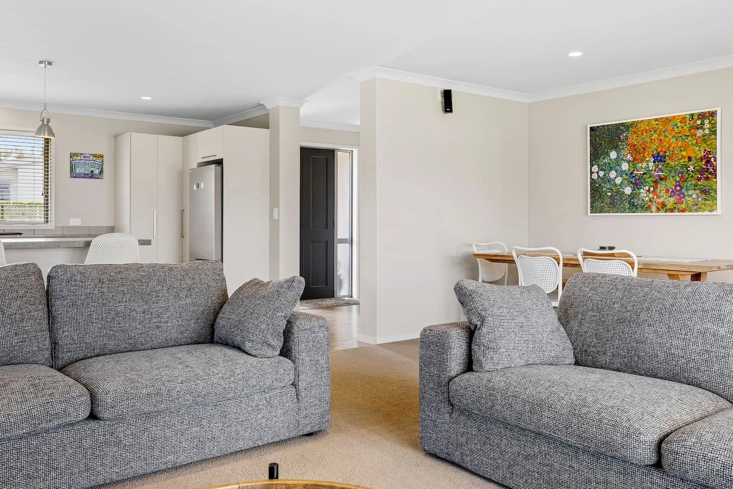Open living room with gray sofas, a dining table with white chairs, and a kitchen area in the background featuring a refrigerator and a window.