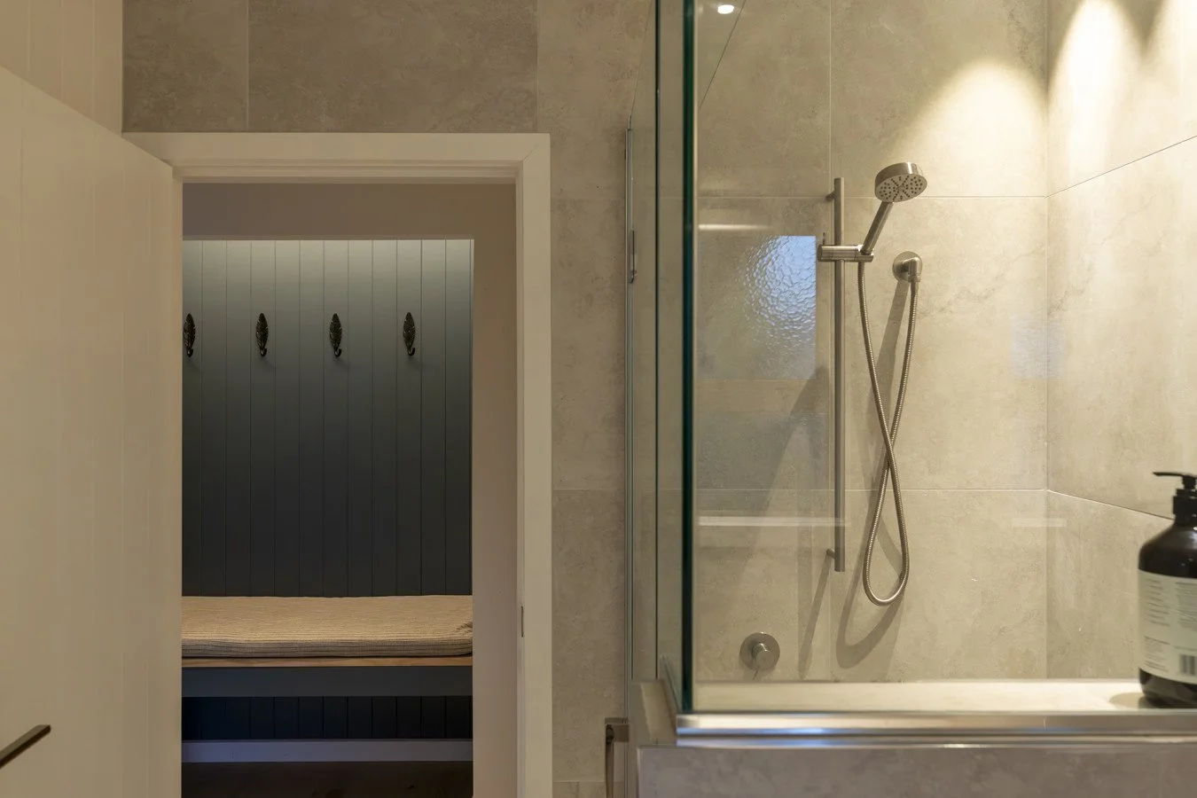 A glass-enclosed shower with a handheld showerhead mounted on a beige tiled wall, and a black bottle on the edge of the bathtub. A doorway leads to a small room with a bench and dark paneling with decorative hooks.