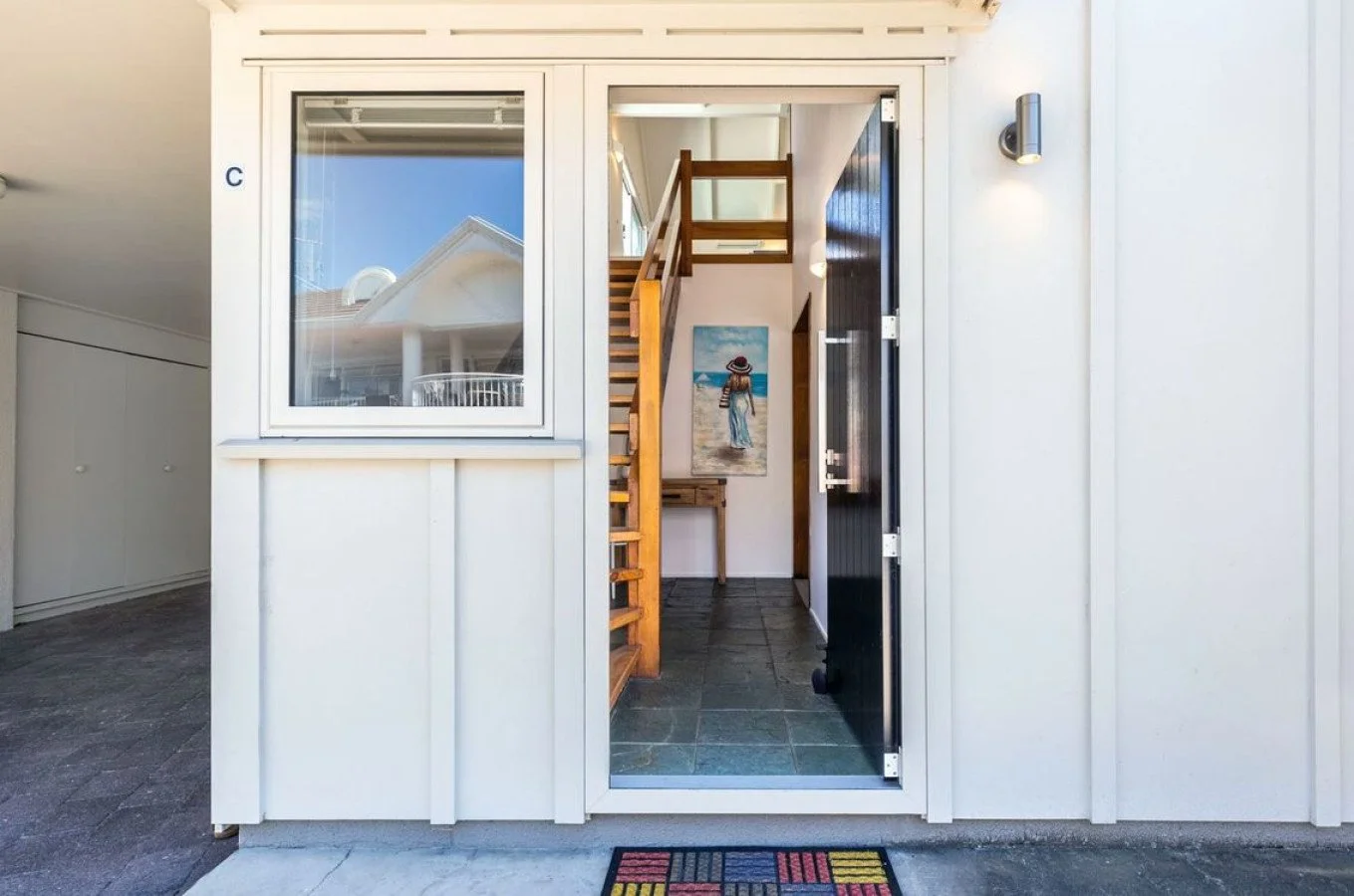 View through a white exterior door revealing a small entryway with a wooden staircase and a painting of a woman on the beach.