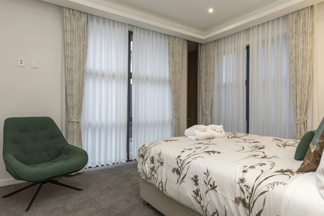 Modern hotel bedroom with a large bed with floral bedding, a green armchair, and large windows with white curtains and beige drapes.