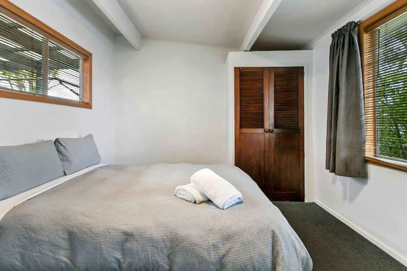 A bedroom with a bed covered in a grey textured blanket and two rolled white towels on top. There are two grey pillows on the bed, a window with blinds on the left, and a window with curtains on the right. A wooden closet with louvered doors is at th