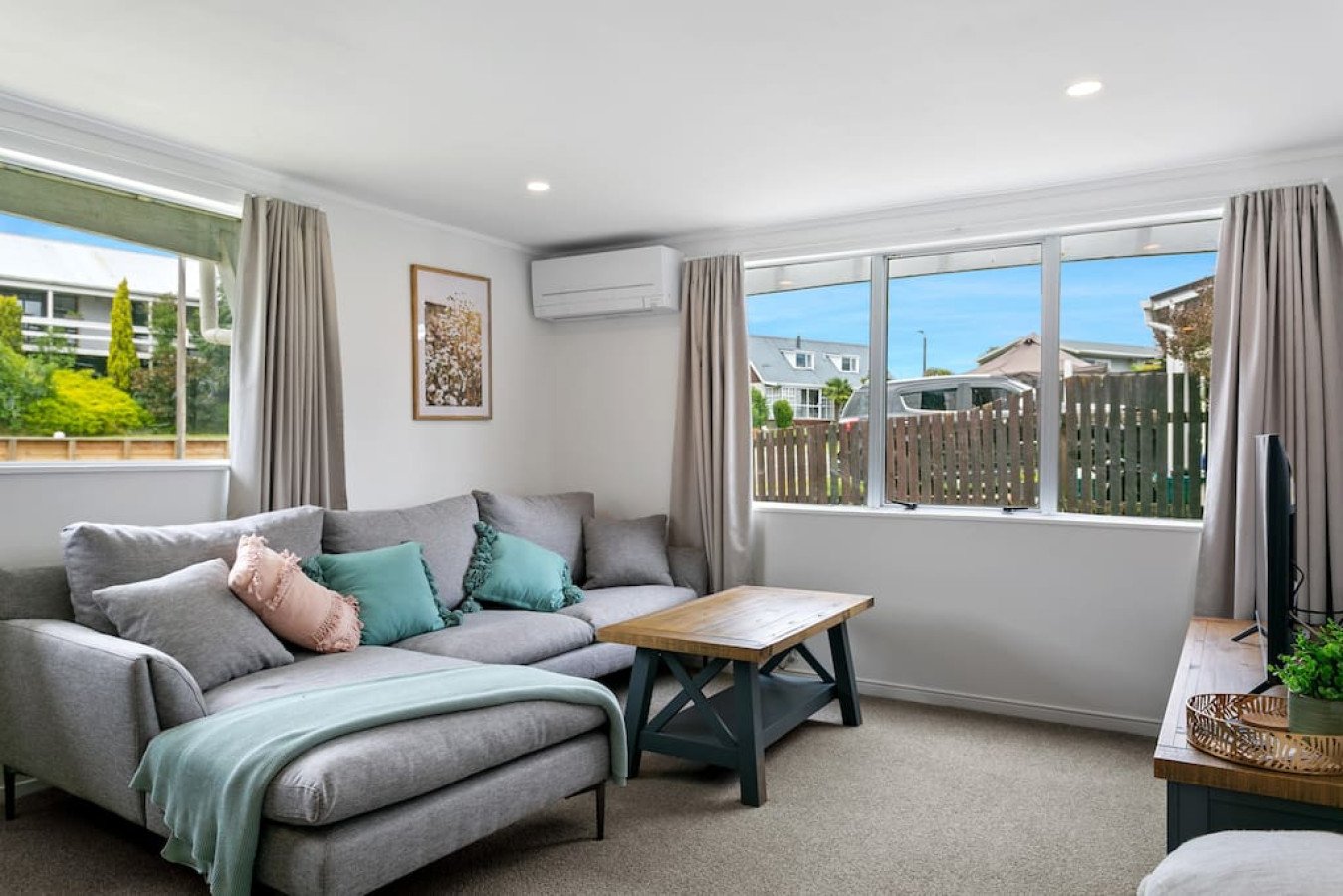 Living room with gray sectional sofa, pastel pillows, wooden coffee table, large windows with garden view, curtains, wall art, air conditioning unit, and a television.