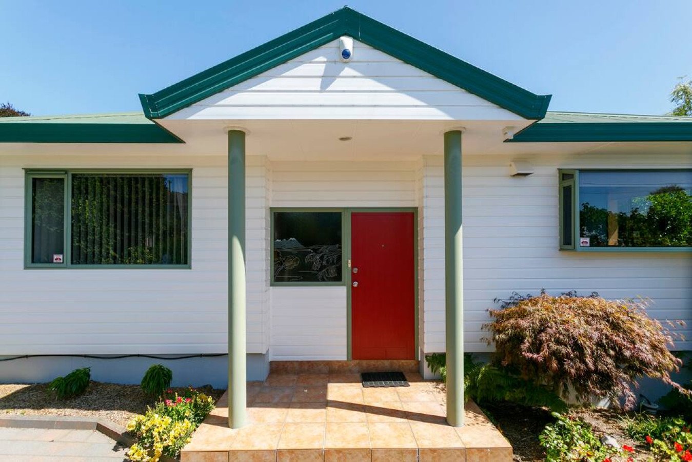Front view of a house with a red door, green trim, and a small porch with two columns. There are windows on either side of the door, and a garden with bushes and flowers in front.