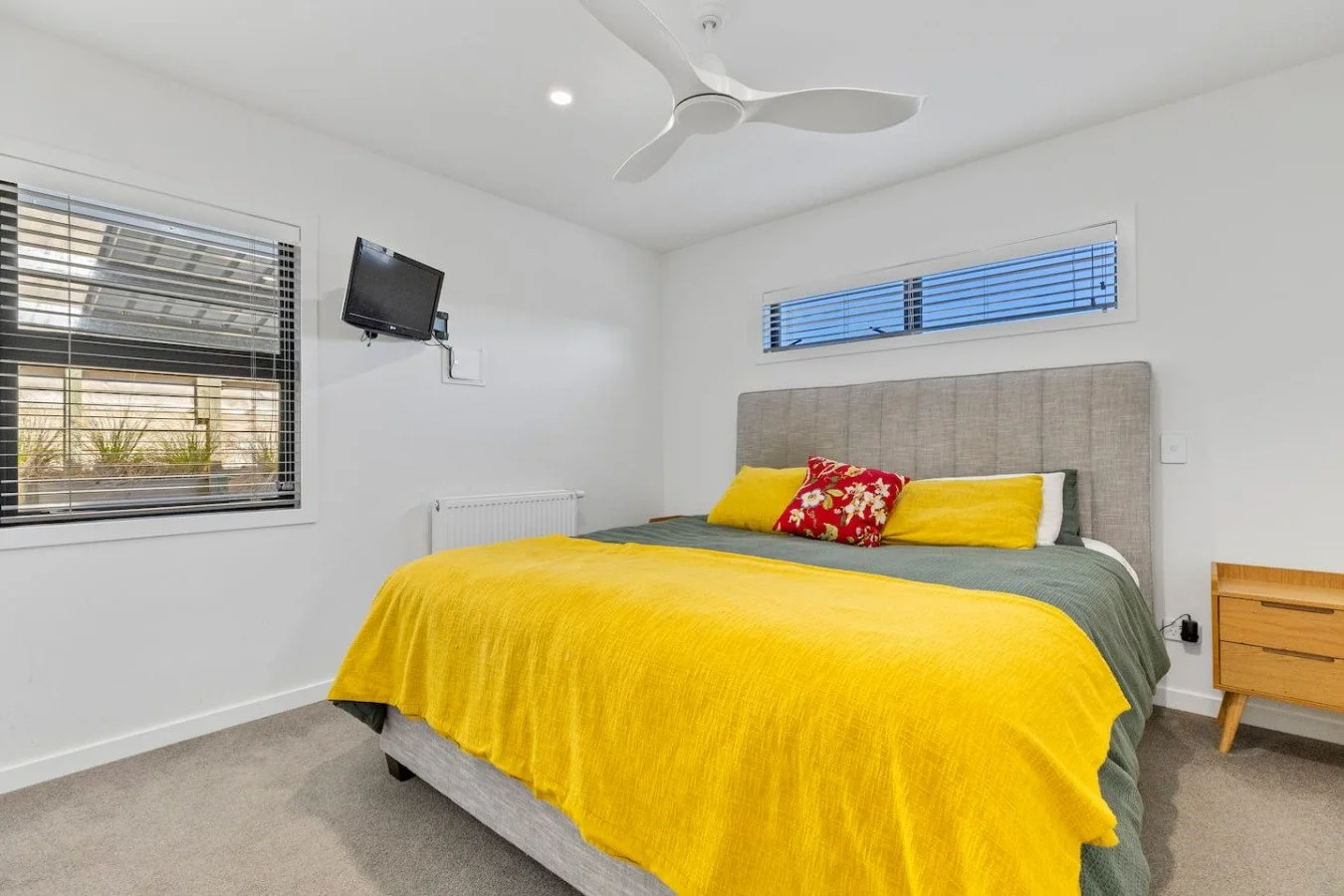 Modern bedroom with a bed covered in a yellow blanket, green sheets, and decorative pillows, a window with blinds, a wall-mounted TV, and a wooden nightstand.