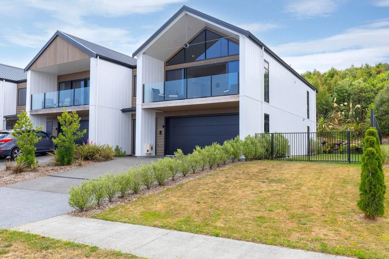 Modern two-story house with white exterior, black garage door, and large glass balcony, surrounded by a small lawn, bushes, and trees, with a hillside and blue sky in the background.