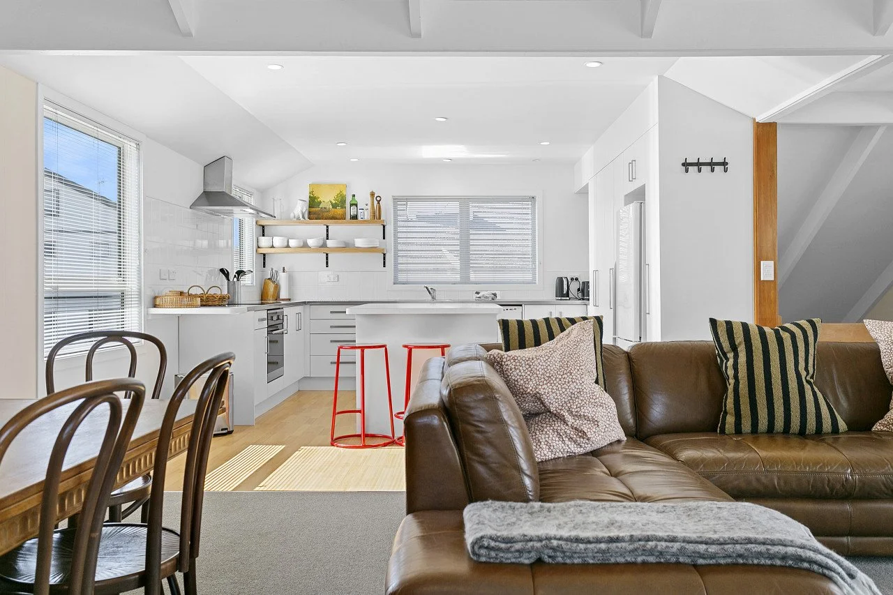 Open-concept living room and kitchen with brown leather sofa, patterned pillows, dining table with chairs, white kitchen with open shelving, windows with blinds, and red barstools at kitchen island.