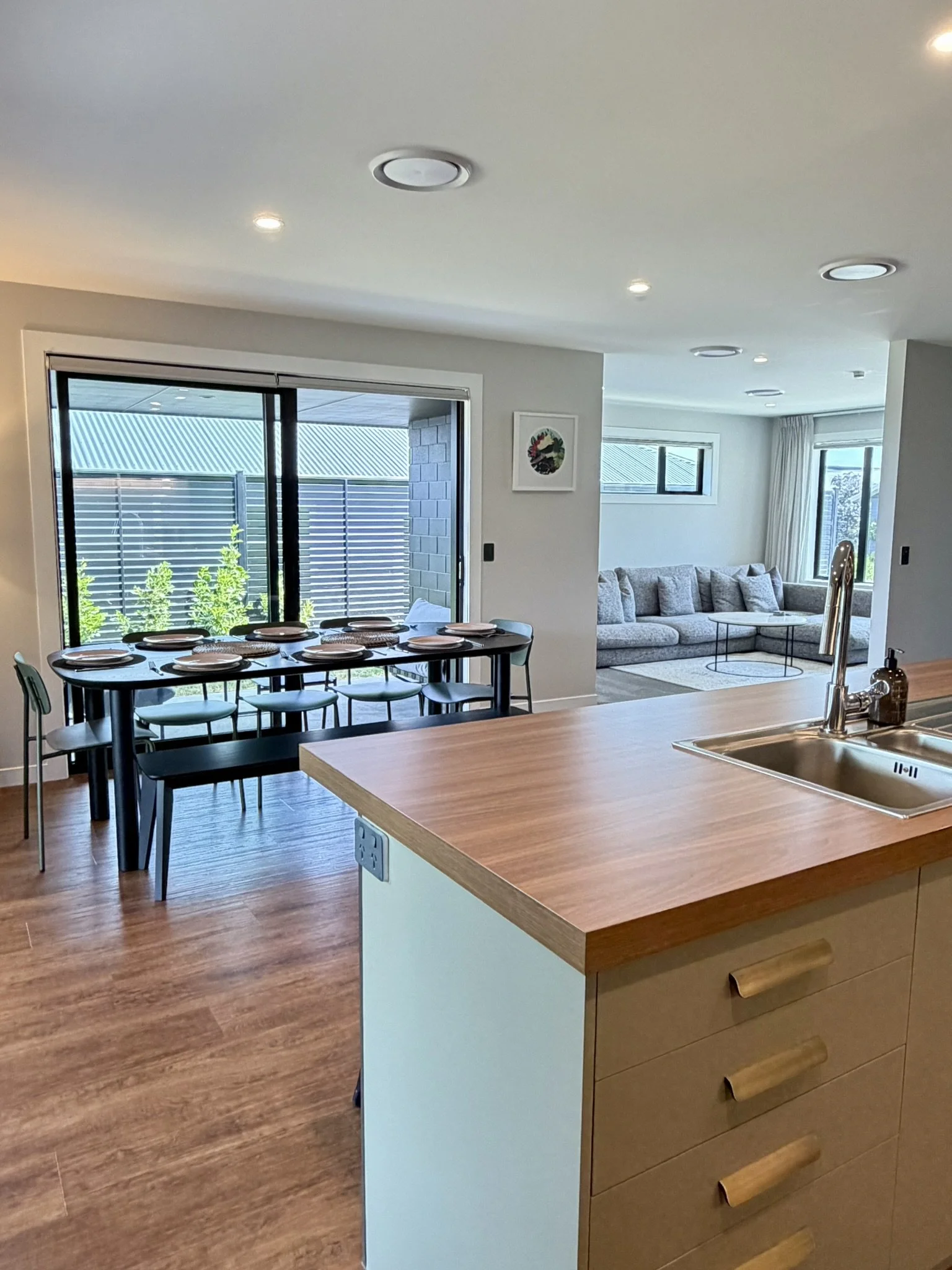 Open-concept kitchen and dining area with a wooden island, a black dining table set for eight, sliding glass doors to a patio, and a living room with a gray sectional sofa and windows.