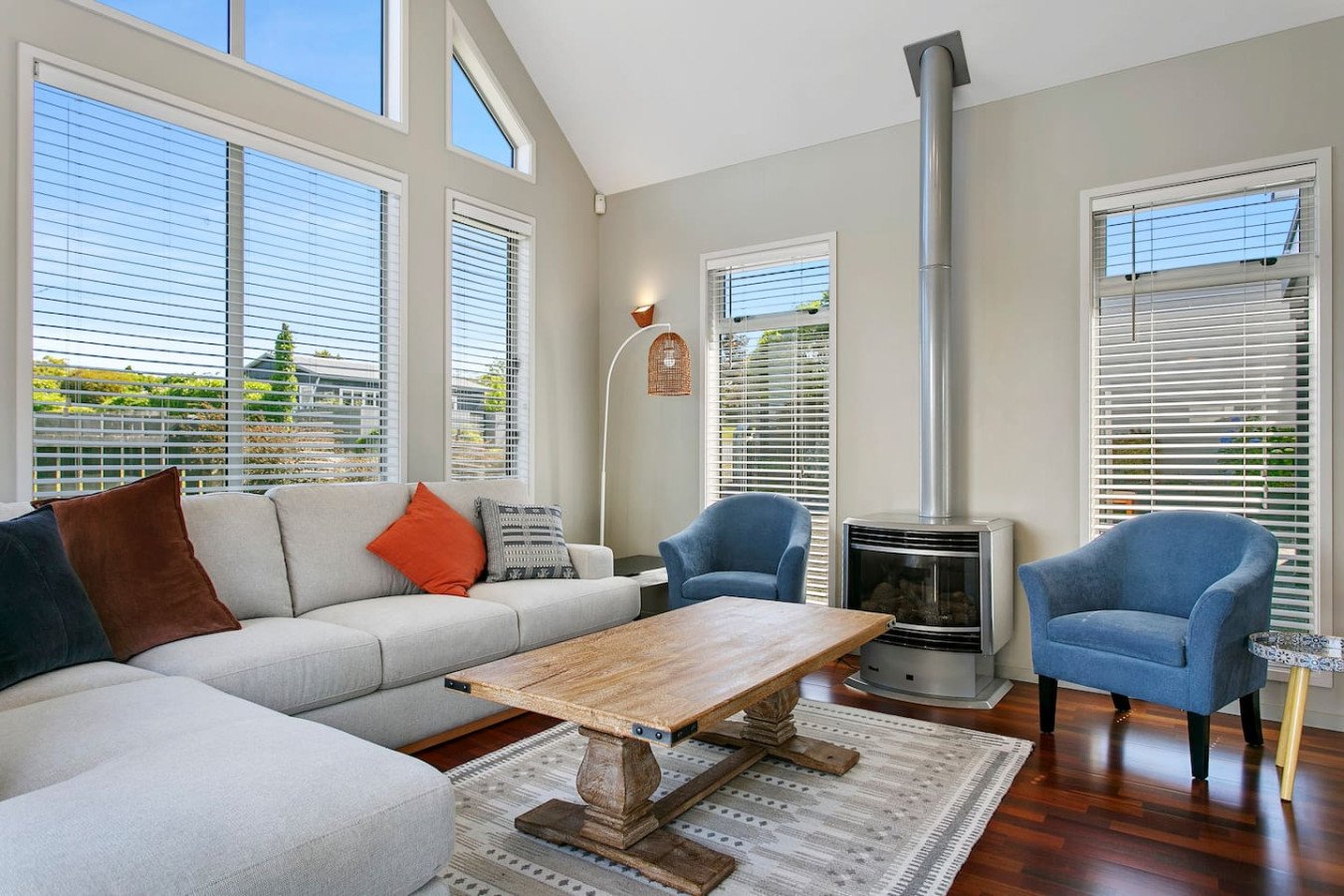 Bright living room with large windows, beige sofa with colorful pillows, blue armchairs, a wooden coffee table, a modern fireplace, and hardwood floors.