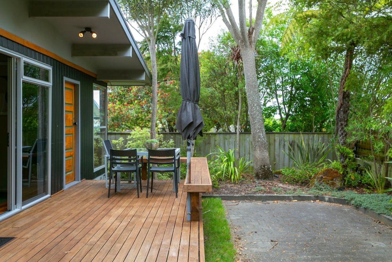 Backyard patio with wooden deck, patio furniture including a table, chairs, and an umbrella, surrounded by trees and plants with a wooden fence.