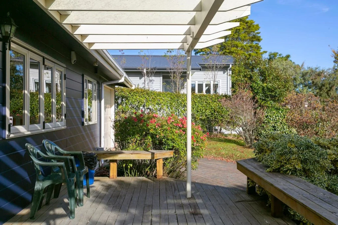 Backyard patio with wooden deck, green plastic chairs, a round table, surrounded by bushes and trees, with a house in the background under a bright blue sky.