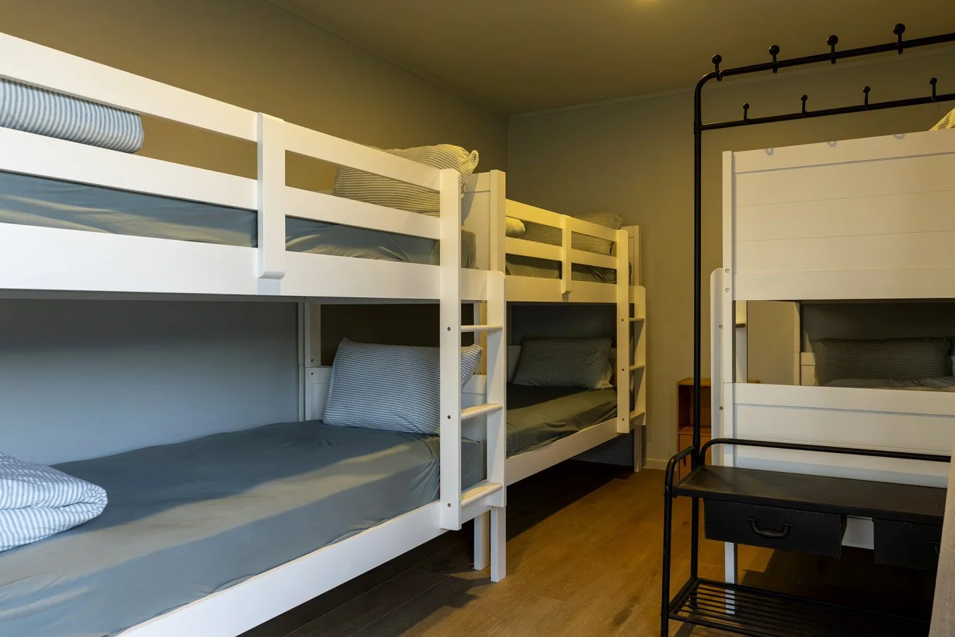 A room with white bunk beds with gray bedding, pillows, and a small black desk with a drawer and a shelf underneath.