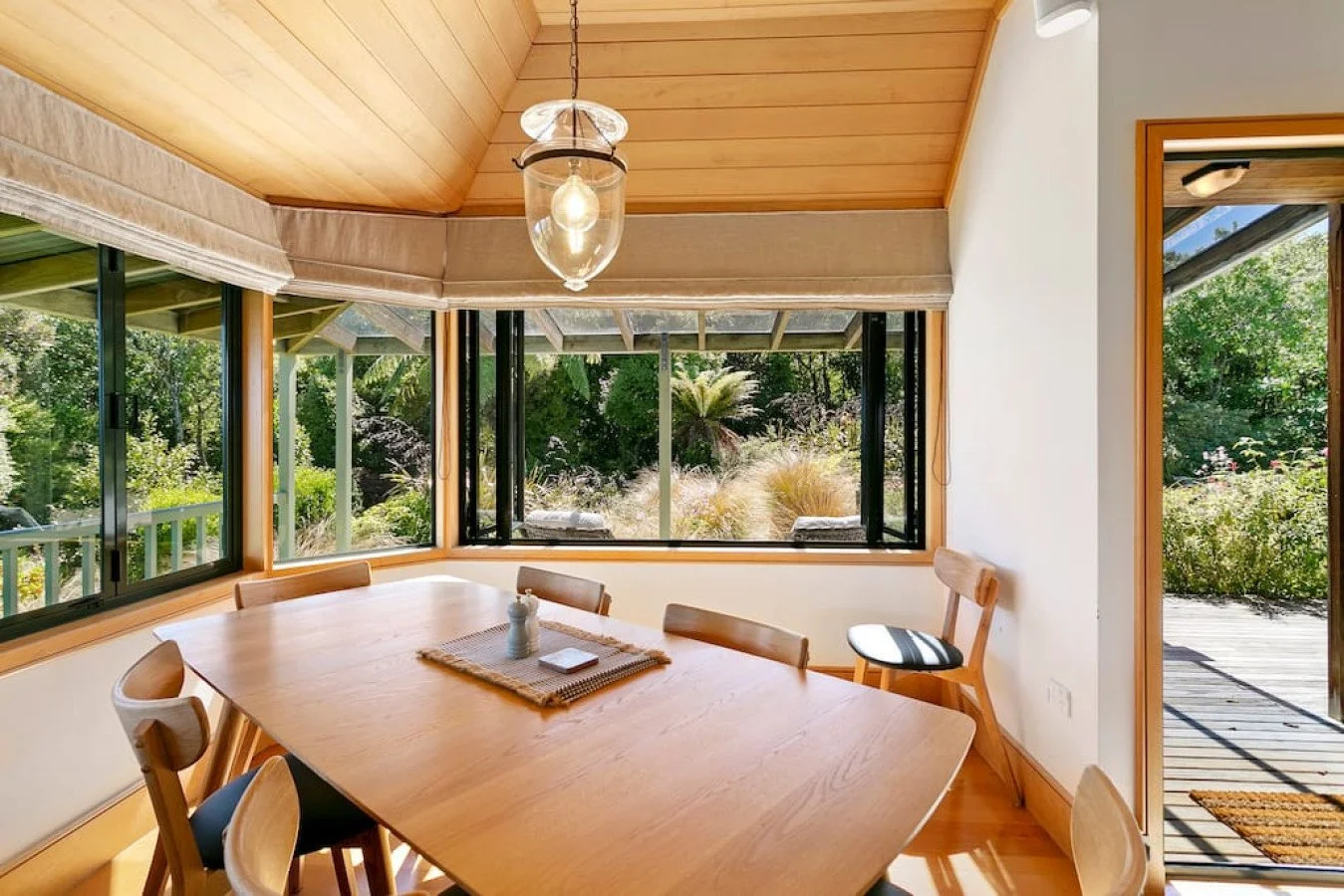 Dining room with wooden table and chairs, large windows showing outdoor greenery, a hanging light fixture, and a sliding glass door leading outside.