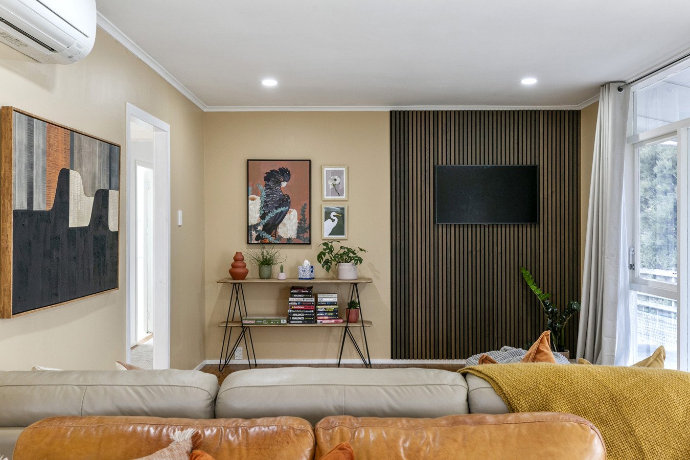 Living room with beige walls, a beige sofa, and a leather armchair. Wall art includes a large abstract painting and smaller bird-themed pieces. A black-slatted accent wall has a mounted TV. Next to the TV is a window with white curtains, and a side t