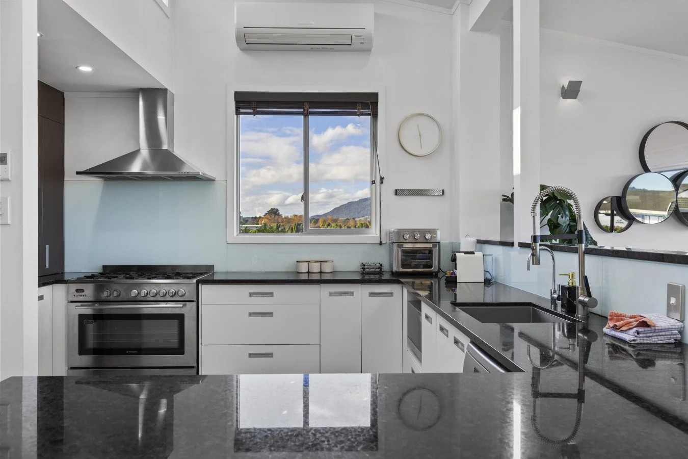 Modern kitchen with white cabinets, black countertops, stainless steel appliances, a window with a view of mountains and blue sky, and decorative round mirrors on the wall.