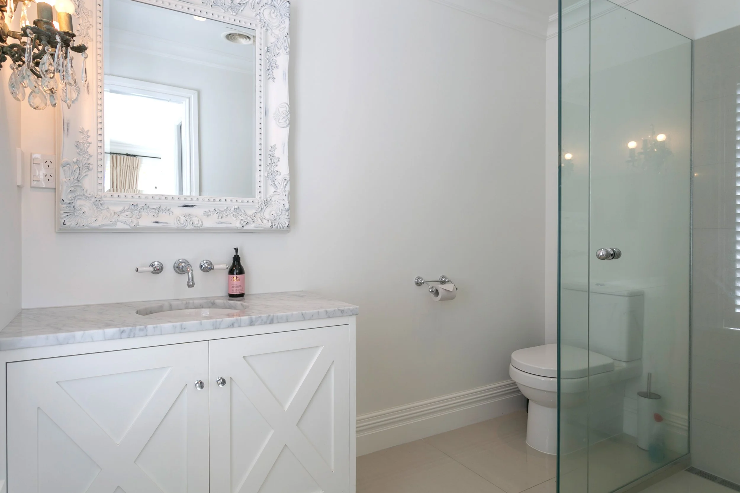 A modern, minimalist bathroom with white walls, a marble countertop vanity with a mirror, a glass-enclosed shower, and a toilet. The vanity features a marble top, and a pink soap dispenser is placed on it. The mirror has an ornate, white frame, and a