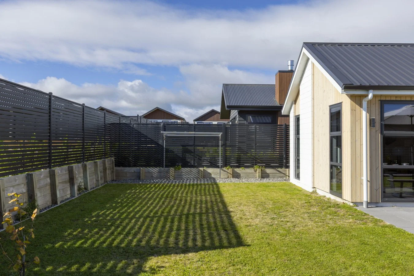 A backyard with a lawn and a modern house, fenced with black horizontal slat fencing, under partly cloudy skies.