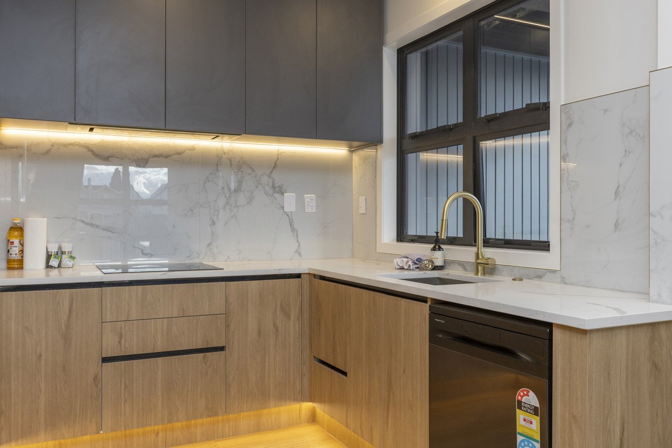 Modern kitchen with wooden cabinets, marble backsplash, black countertop, and a large window above the sink with a gold faucet.