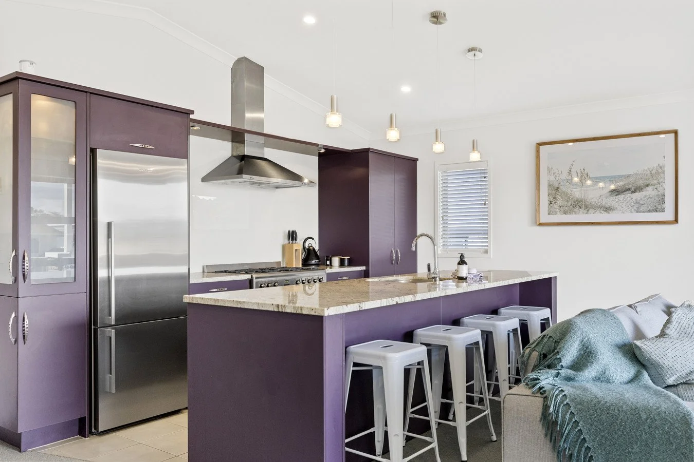 Modern kitchen with purple cabinets, stainless steel refrigerator, and a beige countertop island with four gray bar stools. A sofa with throw pillows and blanket is visible in the living area with a framed artwork on the wall.