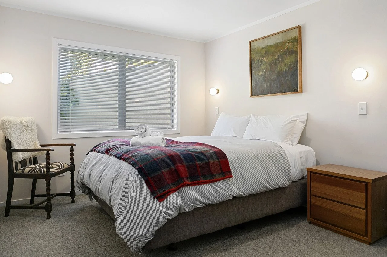 A cozy bedroom with a large bed covered in white linens, a red and plaid blanket, and towel swans. Beside the bed is a wooden nightstand, and a wooden chair with a fluffy blanket draped over it. The room has a large window with blinds, and a framed l