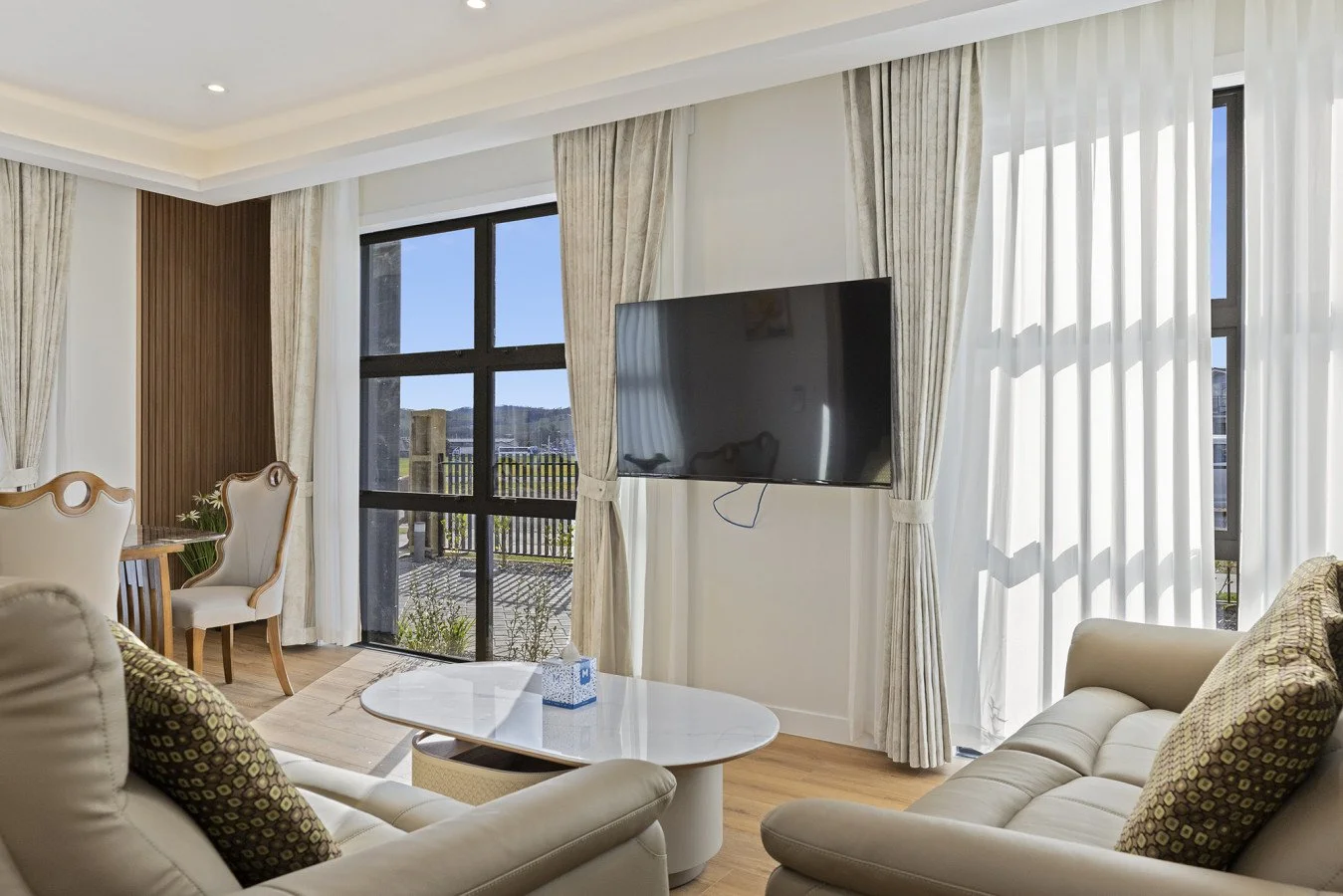 Living room with beige sofa, patterned pillows, white oval coffee table, dining chairs, large windows with white and beige curtains, and a mounted flat-screen TV.