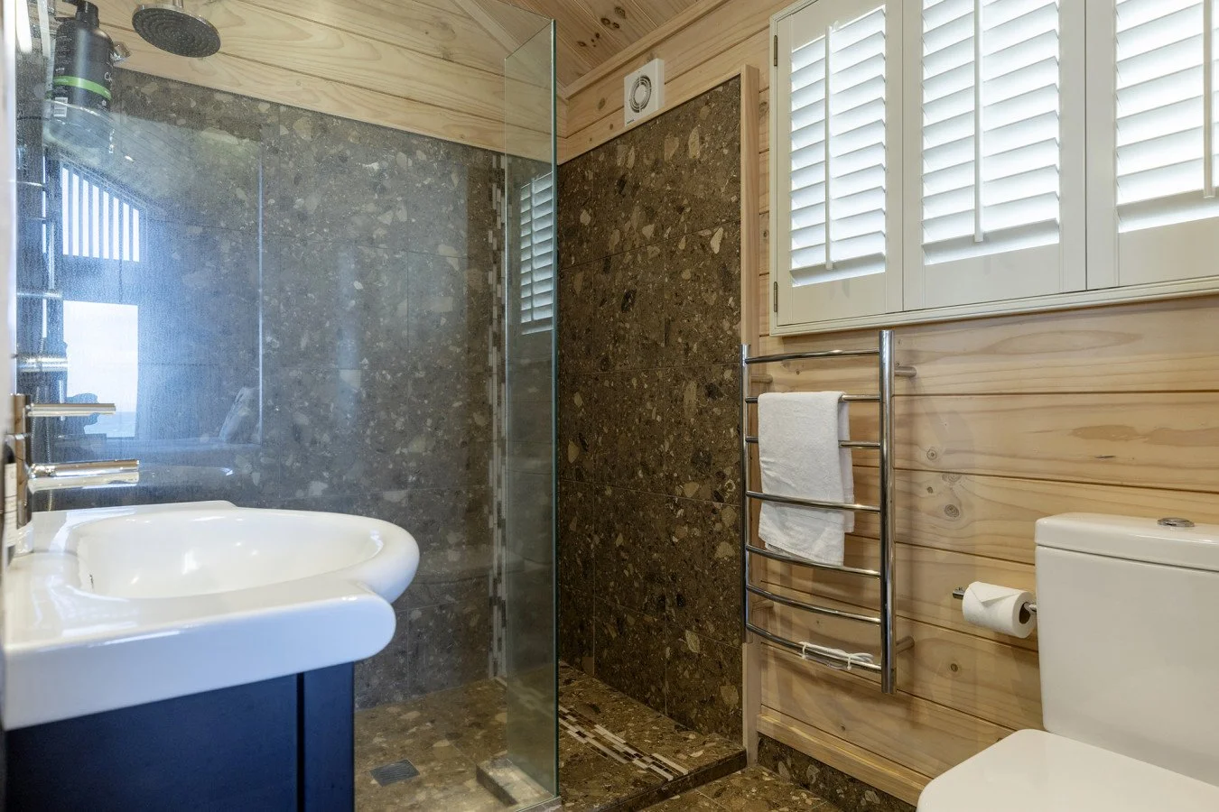 Bathroom with shower, sink, toilet, wooden walls, frosted window, and towel rack.