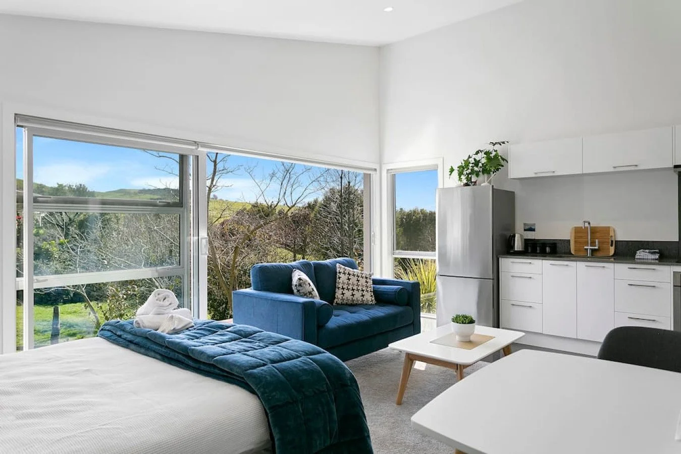 Cozy studio apartment with large windows, a bed with a dark teal quilt, a blue sofa with decorative pillows, a small white coffee table, a kitchenette with white cabinets, a refrigerator, and a small dining table.