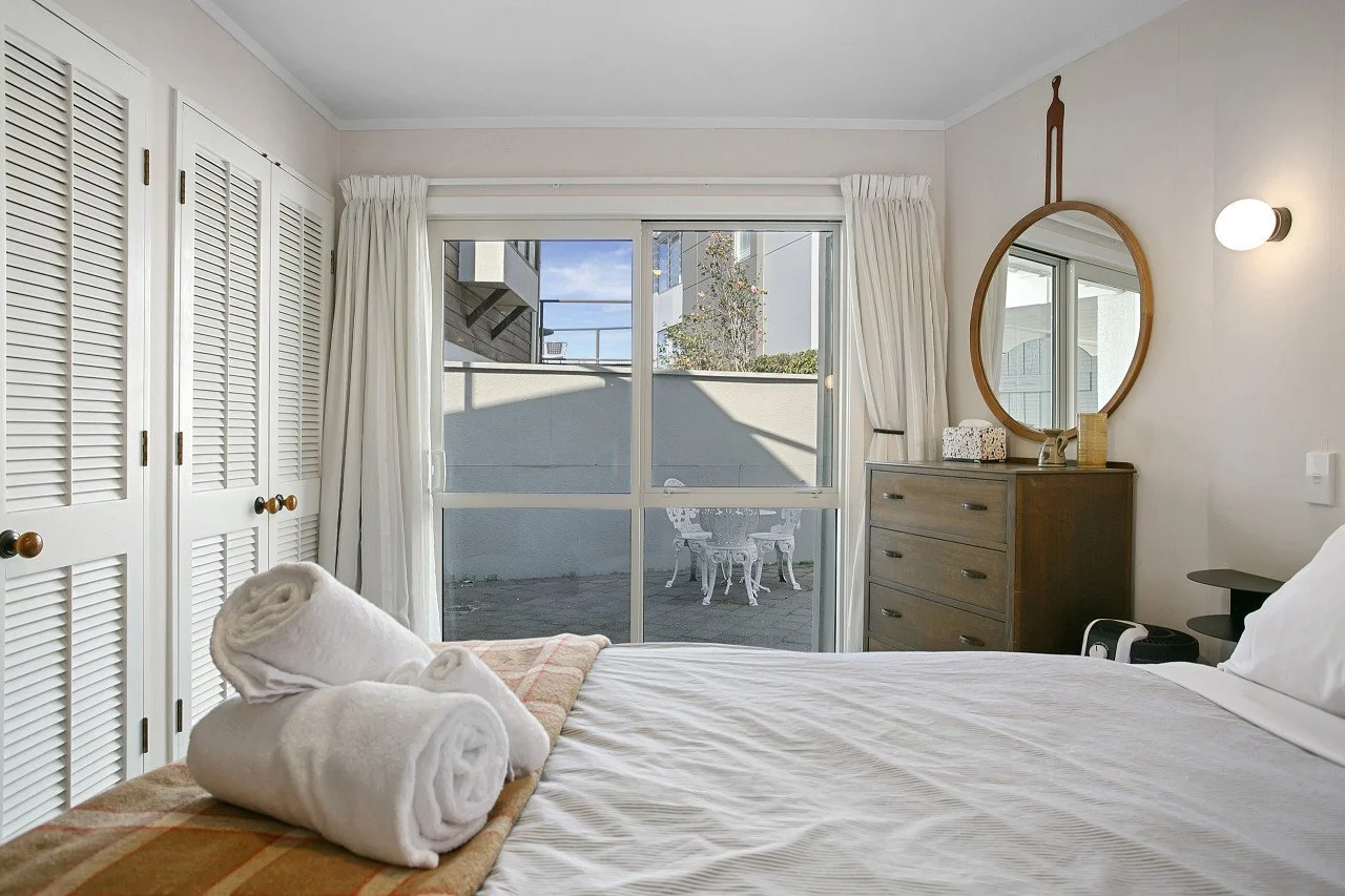 Hotel room with a bed, folded towels, a dresser with a mirror, and a balcony with outdoor furniture visible through sliding glass doors.