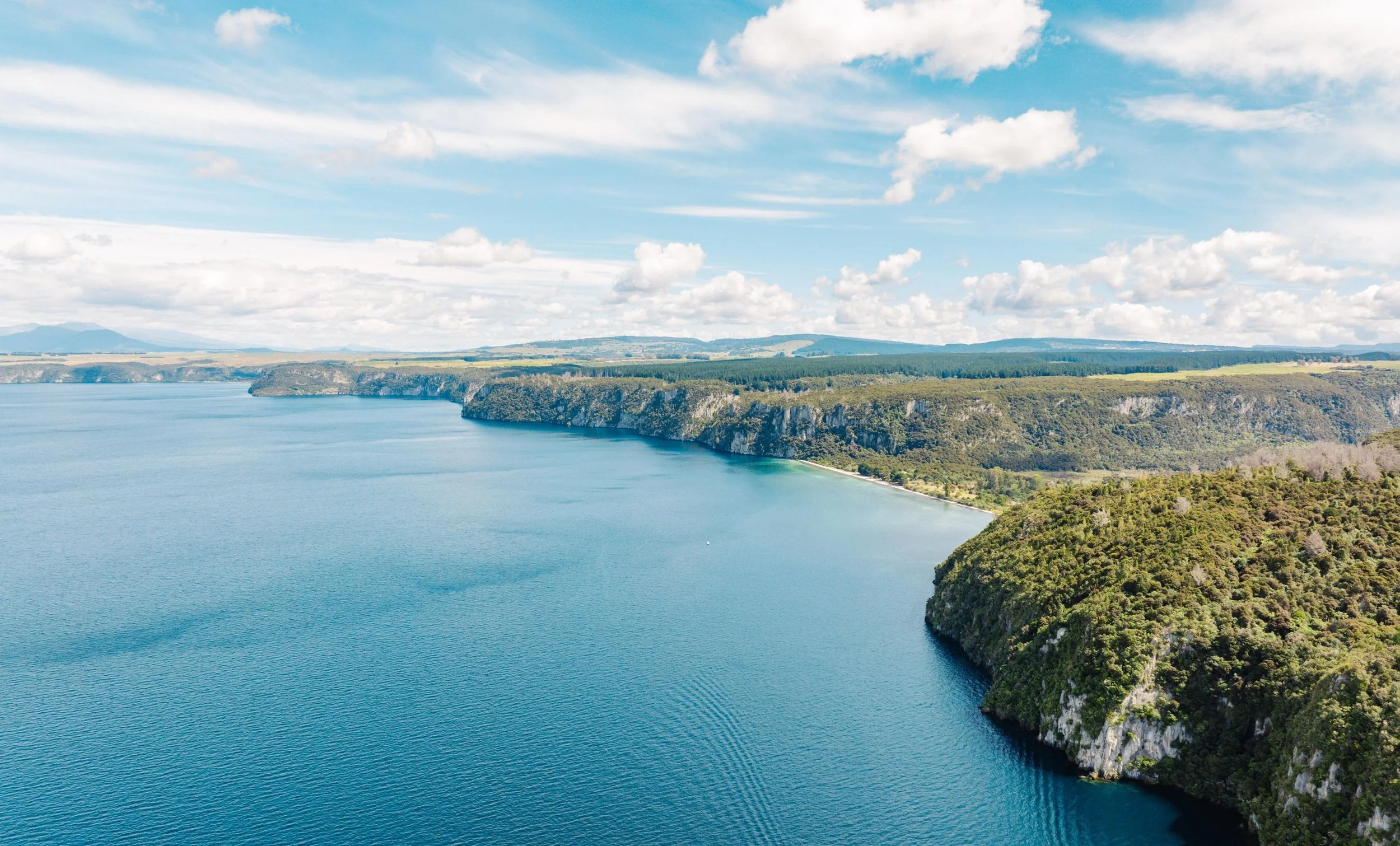 Aerial view of a large body of water with cliffs and greenery along the shoreline, partly cloudy sky.