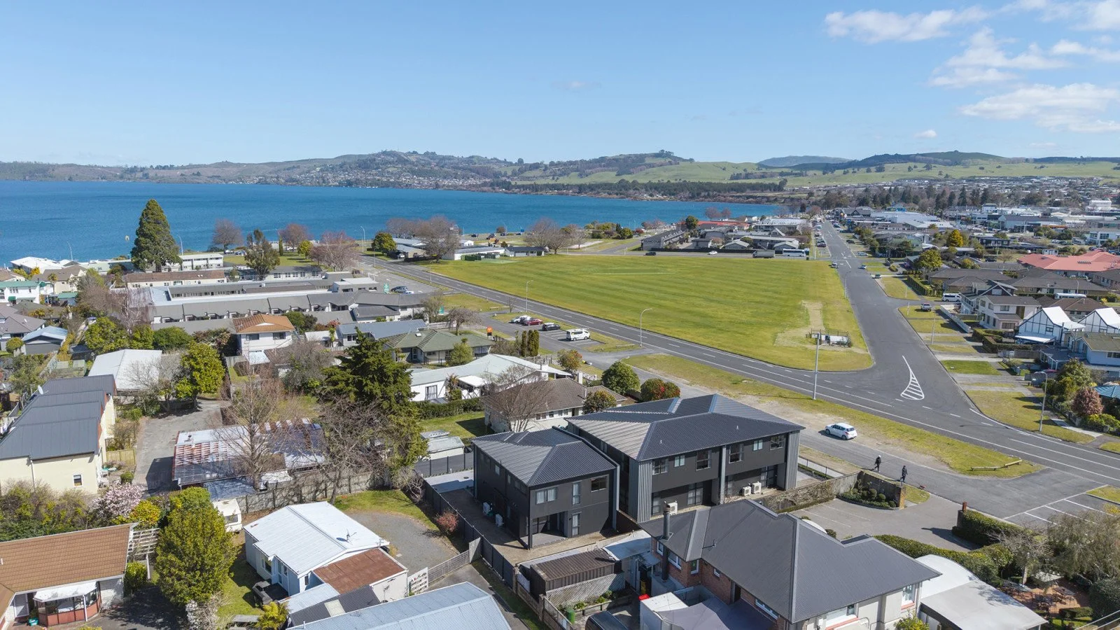 Aerial view of a coastal residential area with houses, a large grassy field, roads, and a body of water in the background under a partly cloudy sky.