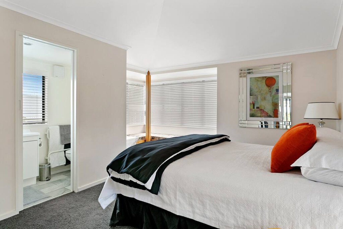 A bedroom with a bed covered in white bedding, orange pillow, and black and white blanket, and a bathroom with a washing machine visible through an open door.